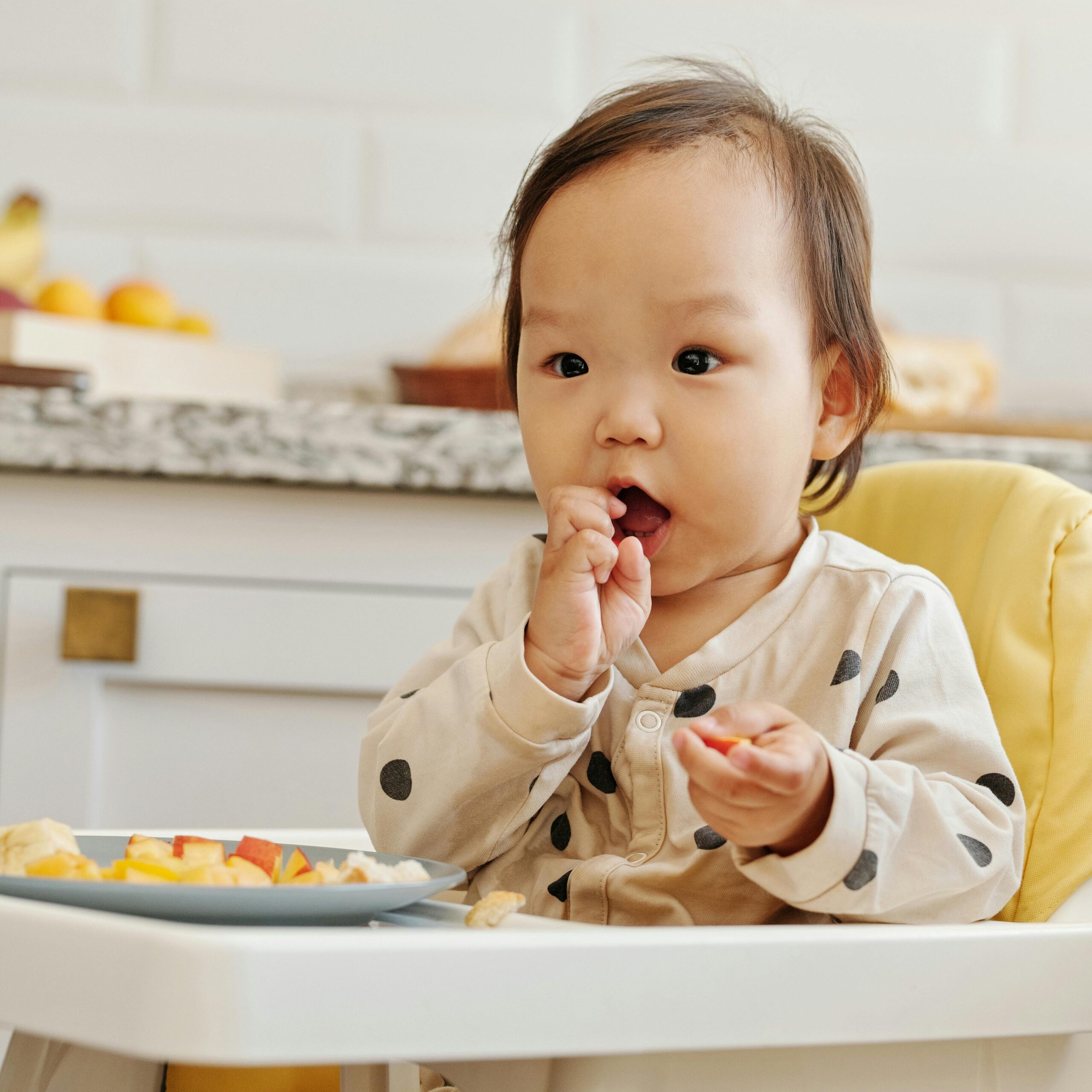 Cute Asian baby in polka dot outfit enjoying healthy fruits in a high chair indoors.